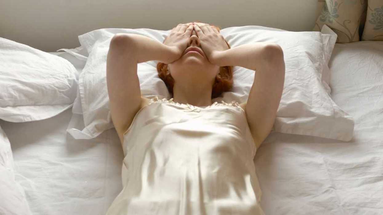 A woman dressed in white lays on her back in a bed with white sheets, she seems frustrated as she holds her hands over her eyes