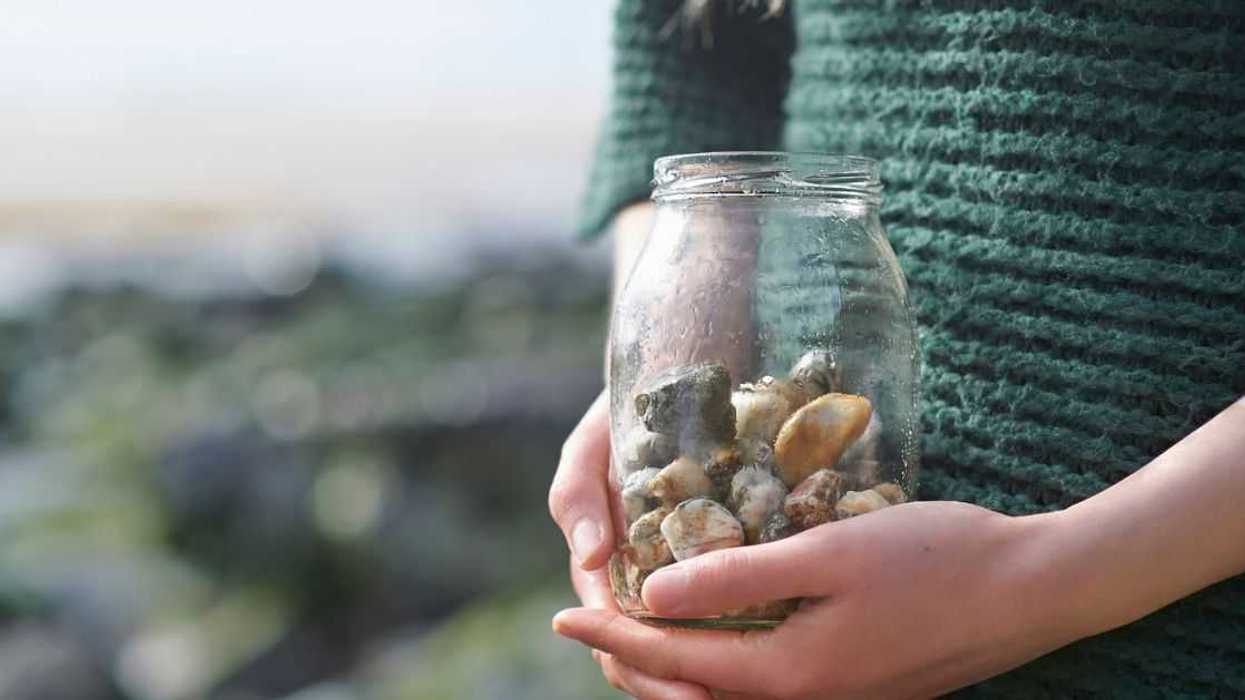 A woman holds a jam jar with stones inside on a rocky beach.