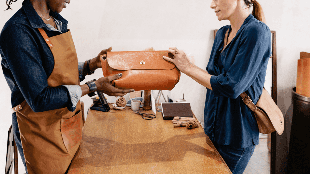 A woman in a leather apron showing a leather handbag to another woman.