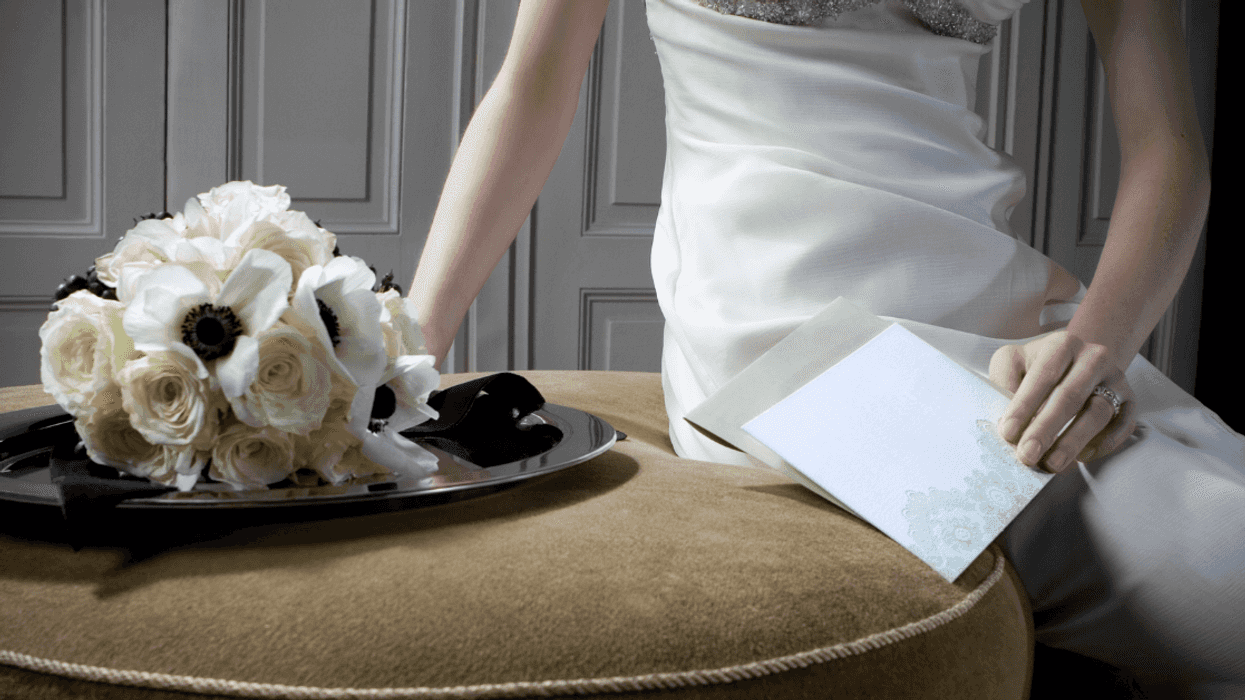 A woman in a wedding dress sitting on table, holding an envelope with a bouquet of flowers.
