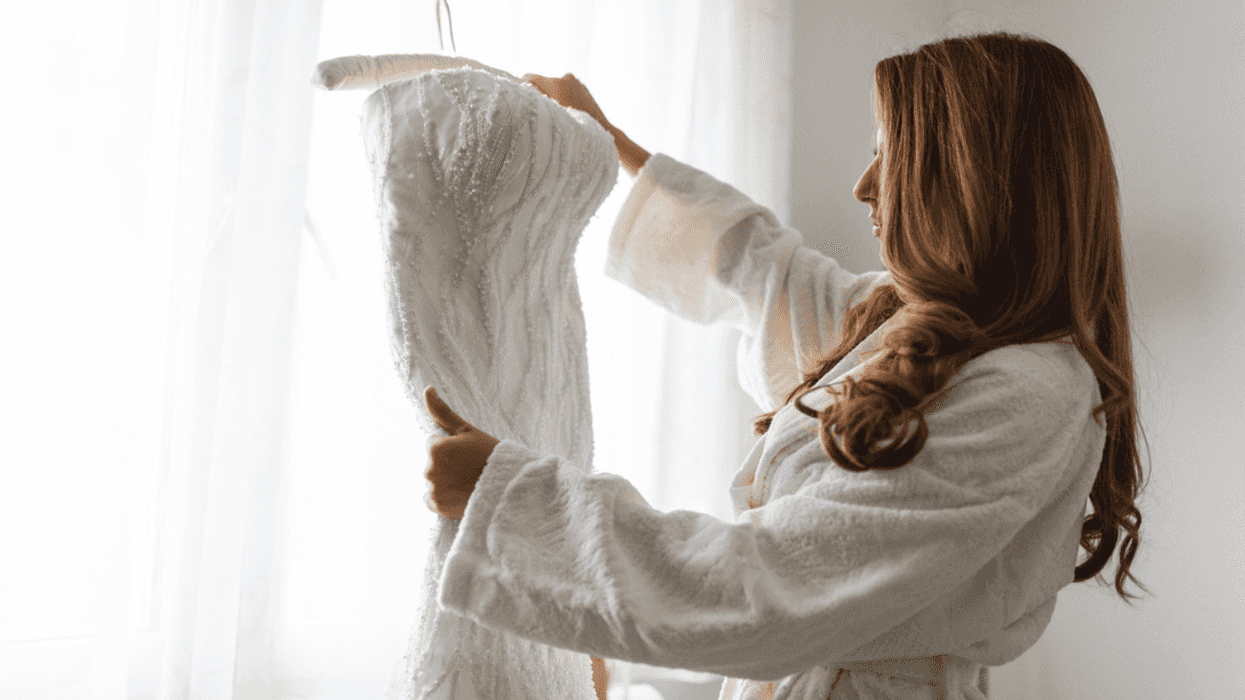 A woman in a white bathroob holding up a wedding dress on a hanger.