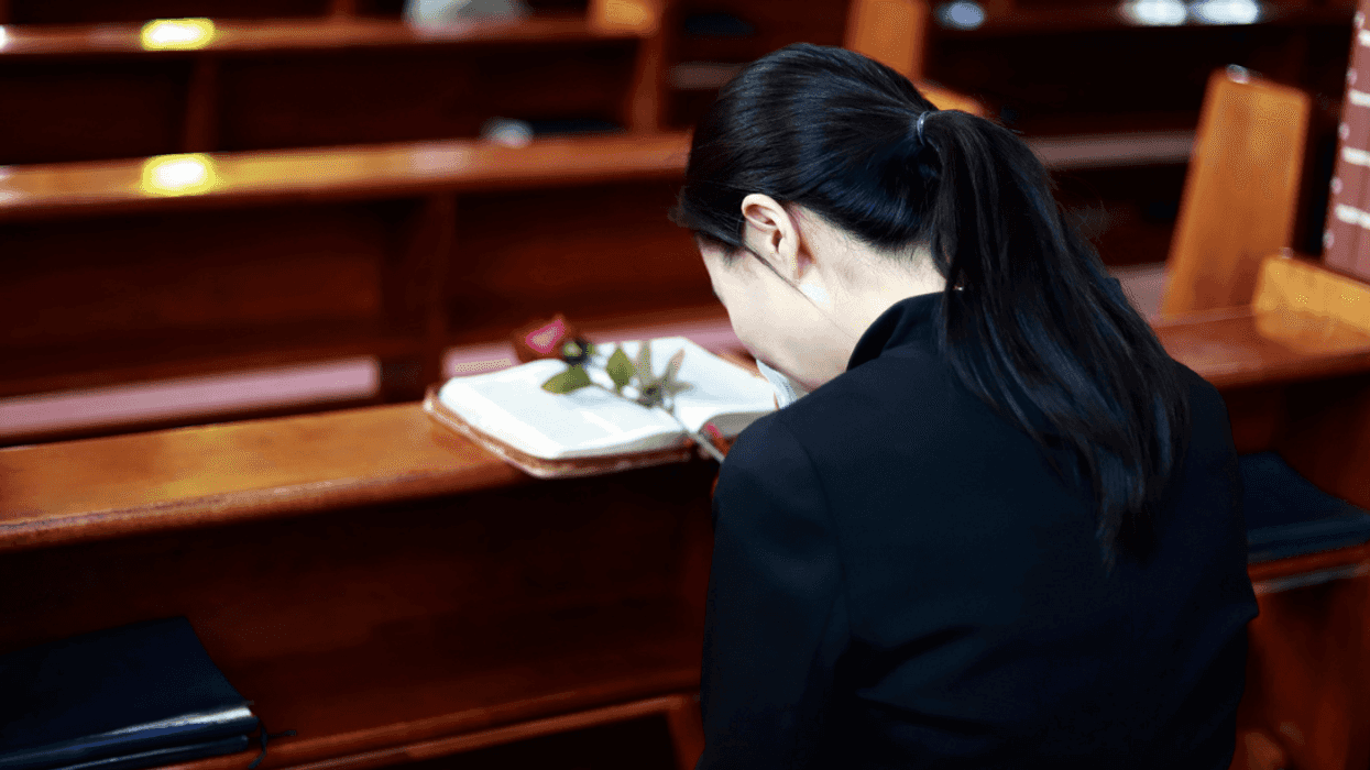 A woman kneeling at a church pew behind an open book with a rose lying on it.