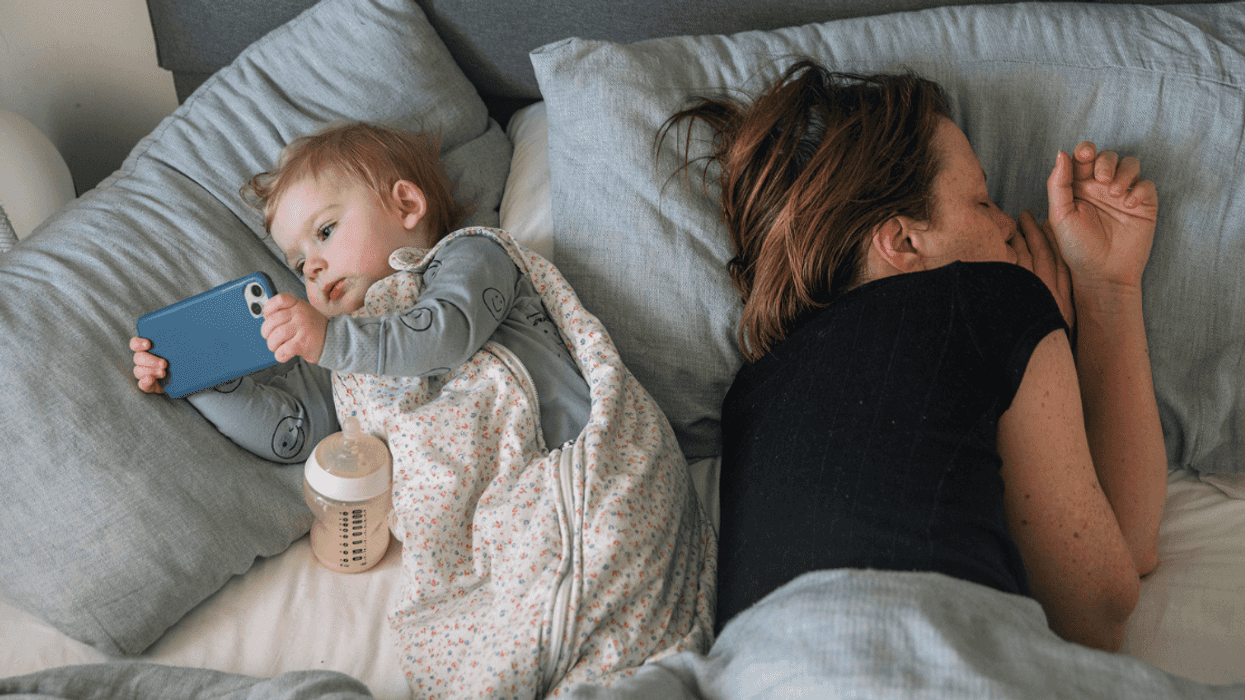 A woman lying down in bed next to a baby holding a smartphone.