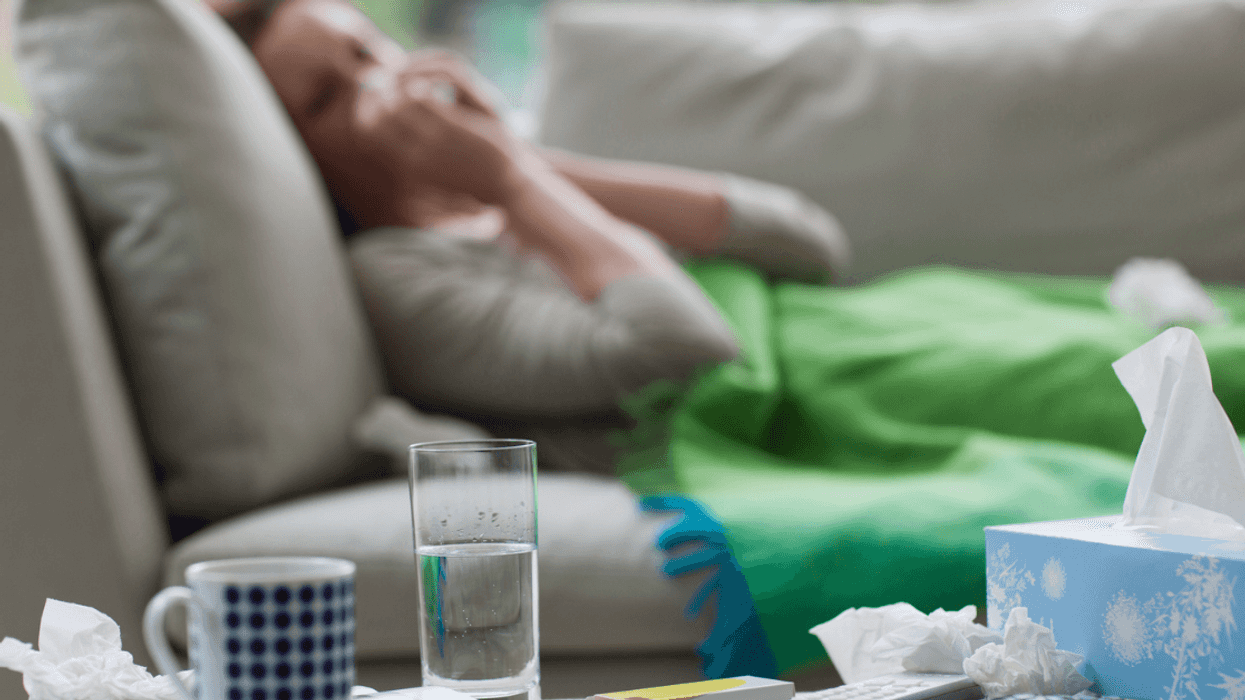 A woman lying on the sofa blowing her nose, with a tissue box, glass of water and coffee mug in front of her.
