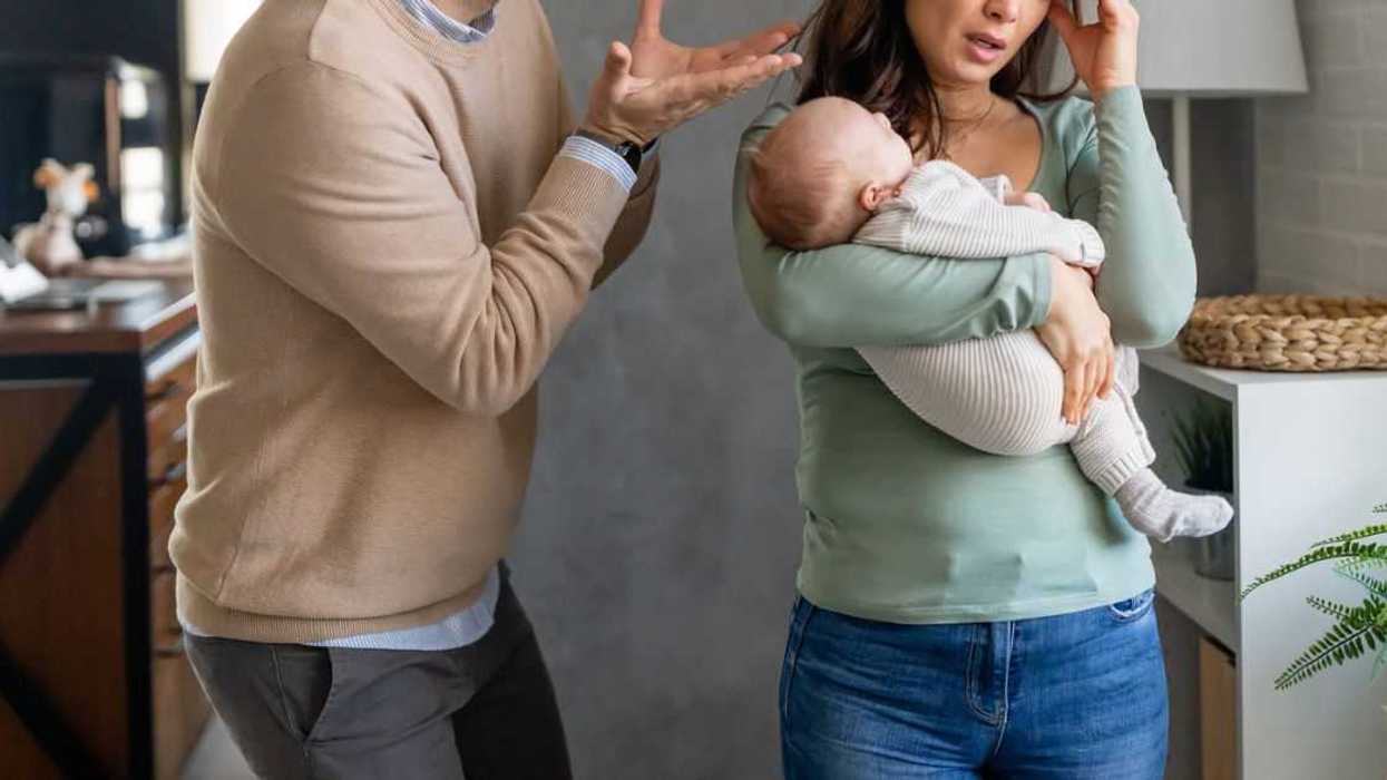 A woman rubs her temple and holds a baby as a man yells at her.