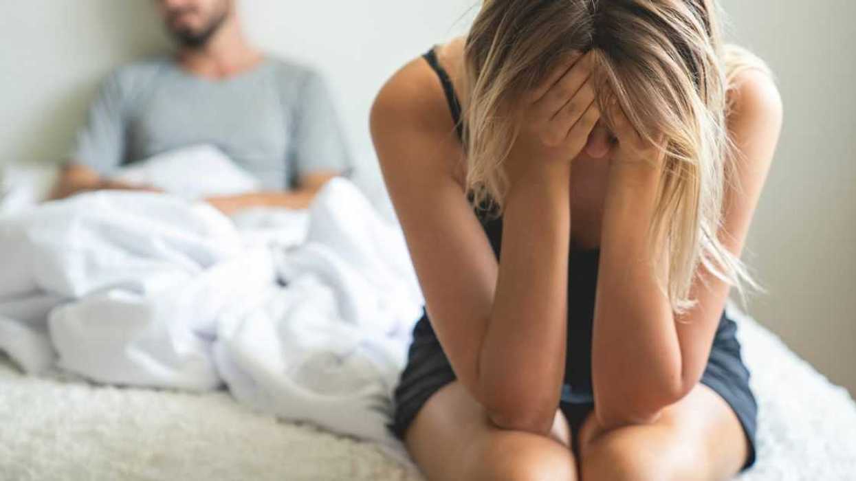 A woman sits on the edge of the bed, man behind her, they've been arguing