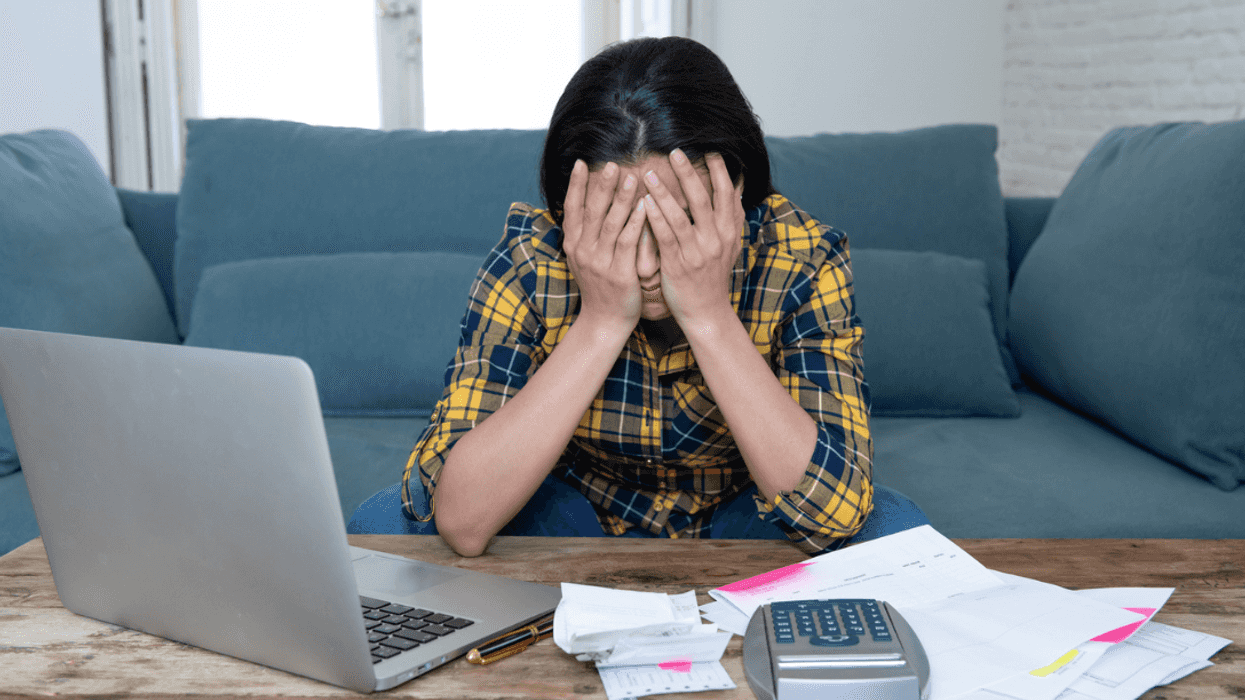 A woman sitting at a coffee table with her head in her hands with a computer, calculator and papers in front of her.