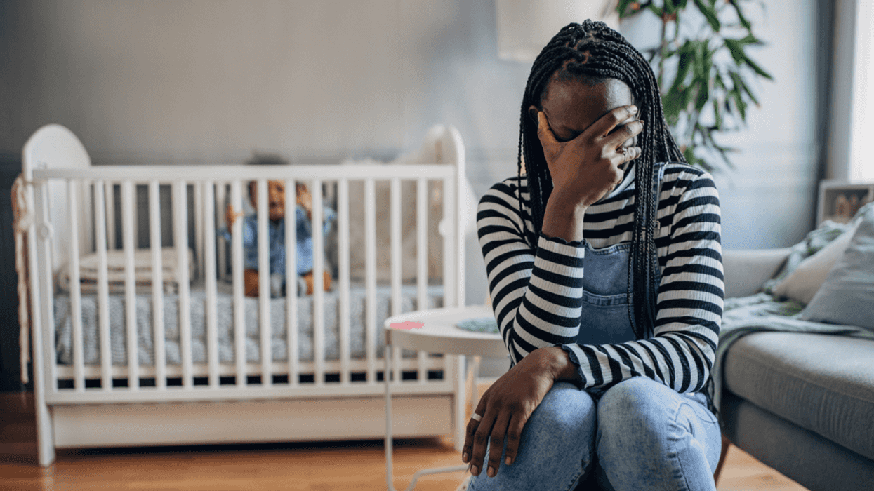 A woman sitting down and crying in front of a crib with an infant in it.