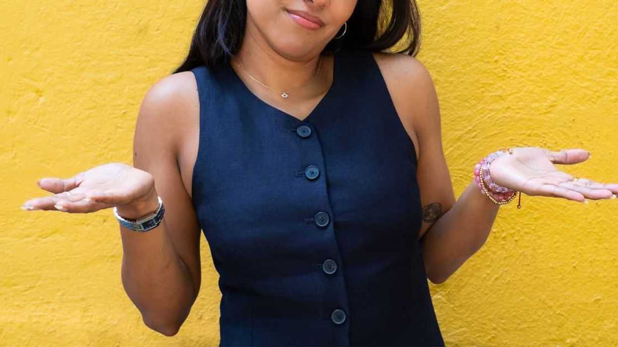 A woman standing against a yellow wall shrugs her hands and head to the side.