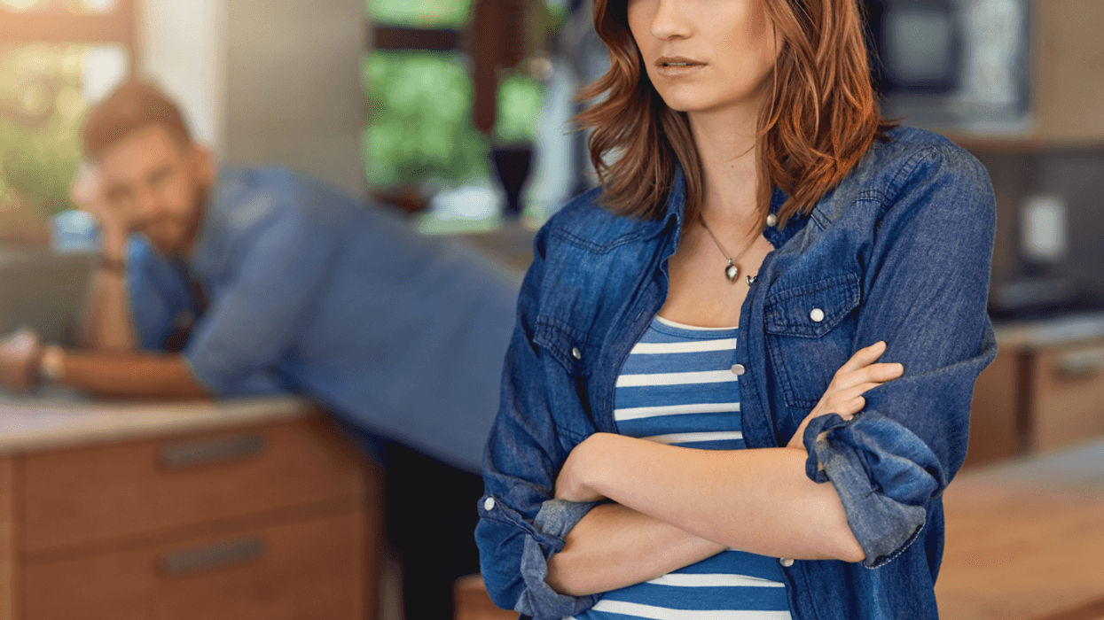 A woman with her arms folded and a man leaning against a counter in the background.