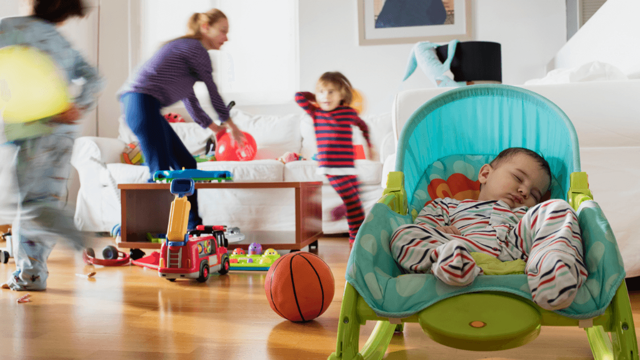 A woman with two children running around her and a baby asleep in a chair.
