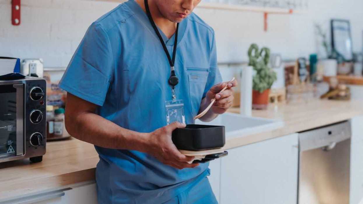 A young adult male nurse leans against the kitchen counter in a hospital break room, eating lunch during a work break.