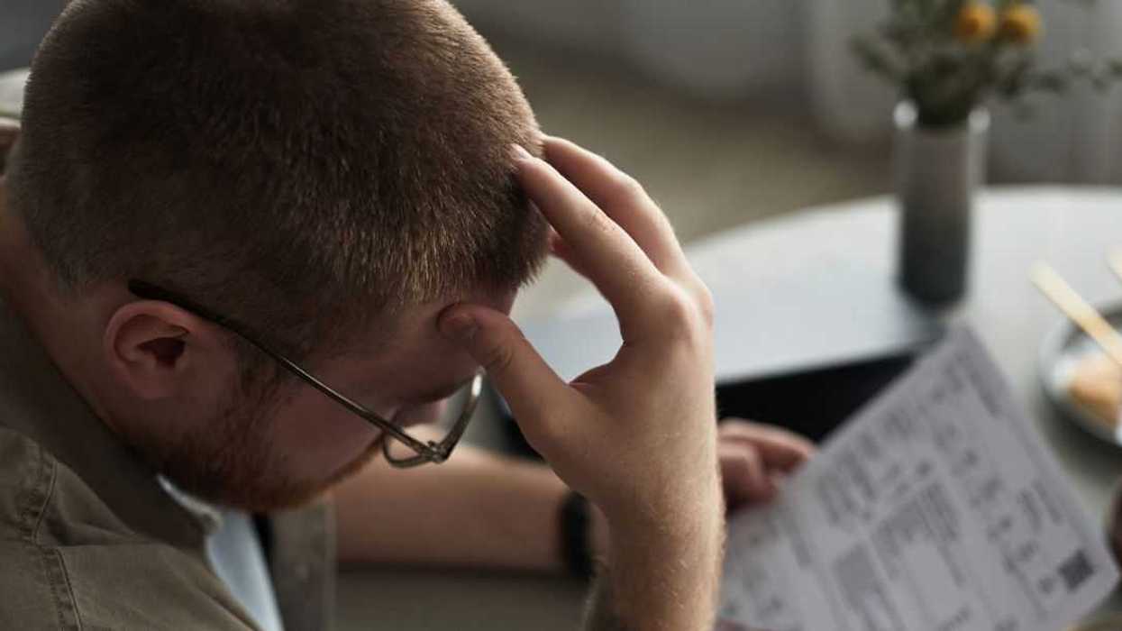A young adult man sits at a table holding overdue bills, looking stressed and frustrated, hand on forehead, financial documents and chopsticks visible on surface.