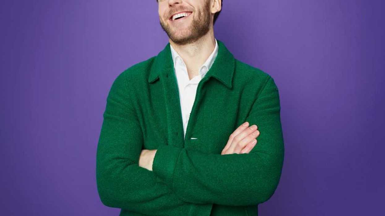 A young, bearded man with his arms folded stands in front of a purple background. He wears a green sweater and he throws in head back in laughter.