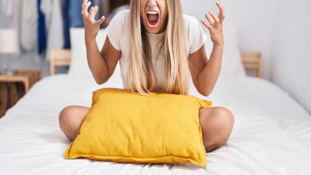 A young blonde woman sitting on the bed, with a pillow, at home, crazy and mad, shouting and yelling with aggressive expression and arms raised.