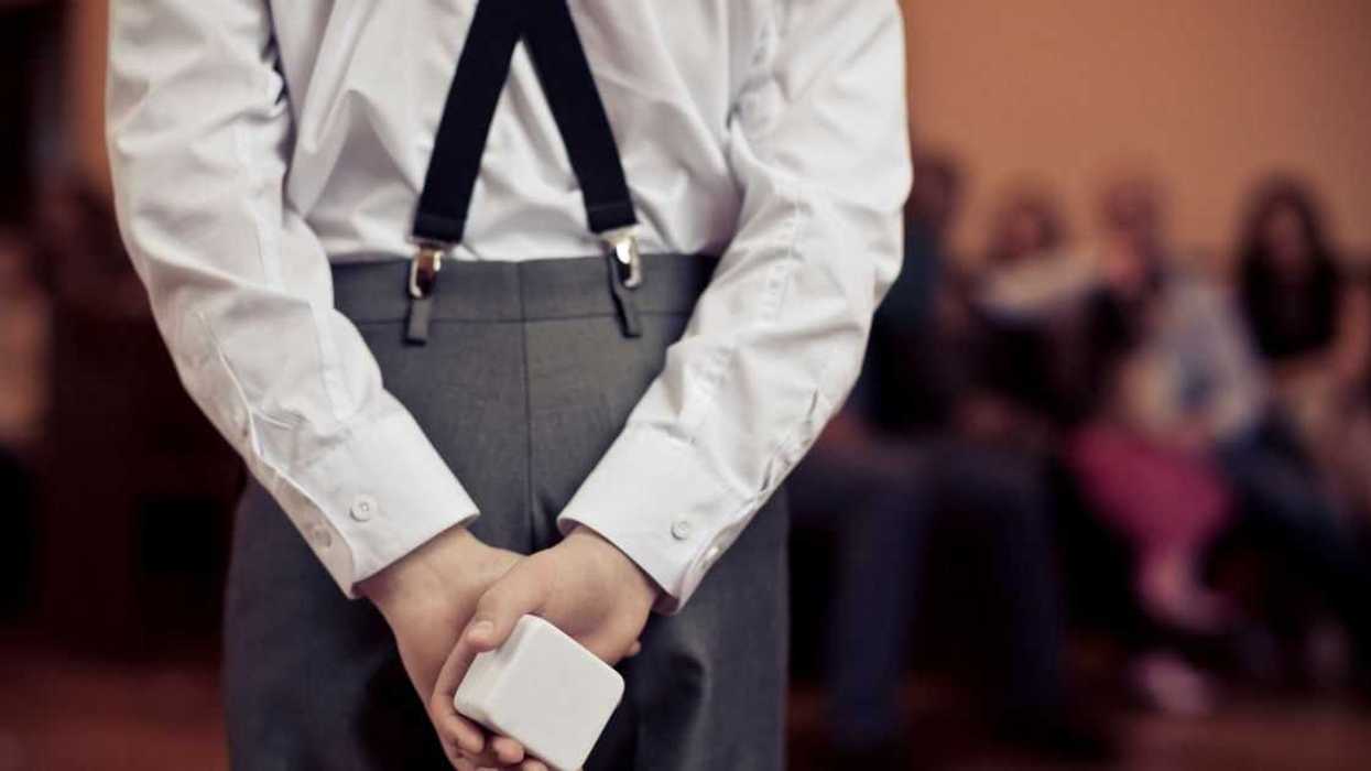 A young boy at a wedding holds a ring in a box behind his back