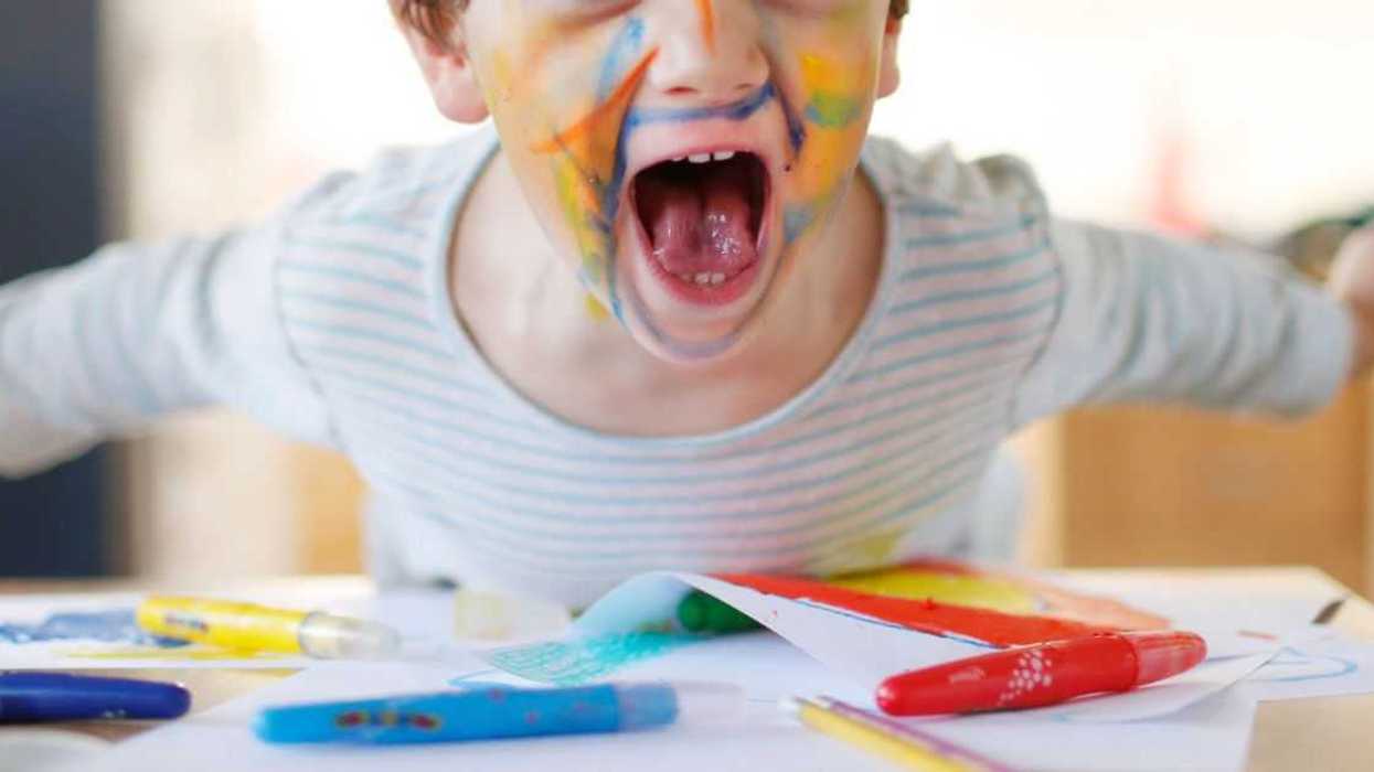 A young child with his face covered in colorful paints yells into the camera