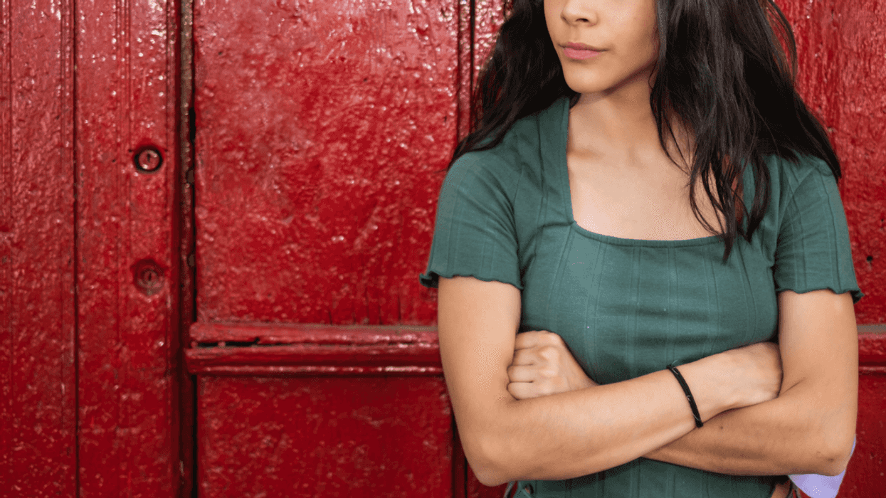 A young girl standing in front of a red door with her arms crossed.