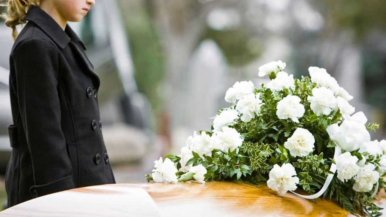 A young girl stands at a coffin in a cemetery. White carnations adorn the casket.