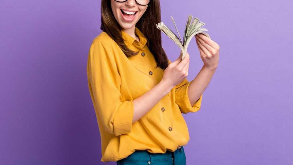 A young, smiling woman fans dollar bills while standing in front of a purple background.