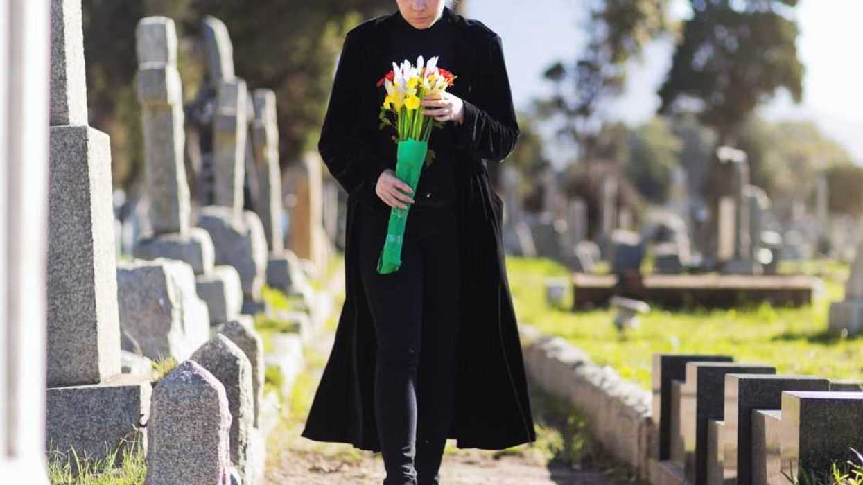 A young woman in black walks through cemetery headstones carrying flowers to the grave of someone she misses.