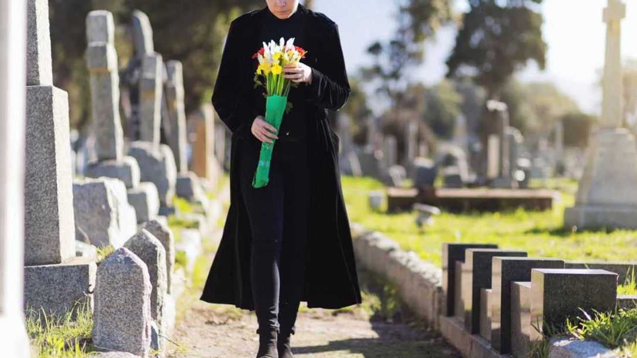 A young woman in black walks through cemetery headstones carrying flowers to the grave of someone she misses.