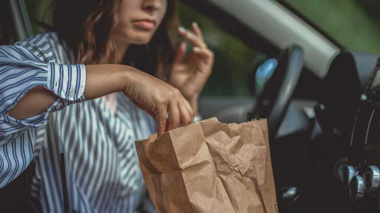 A young woman is reaching for some french fries in a paper bag and she can't wait to start the brunch in her car.