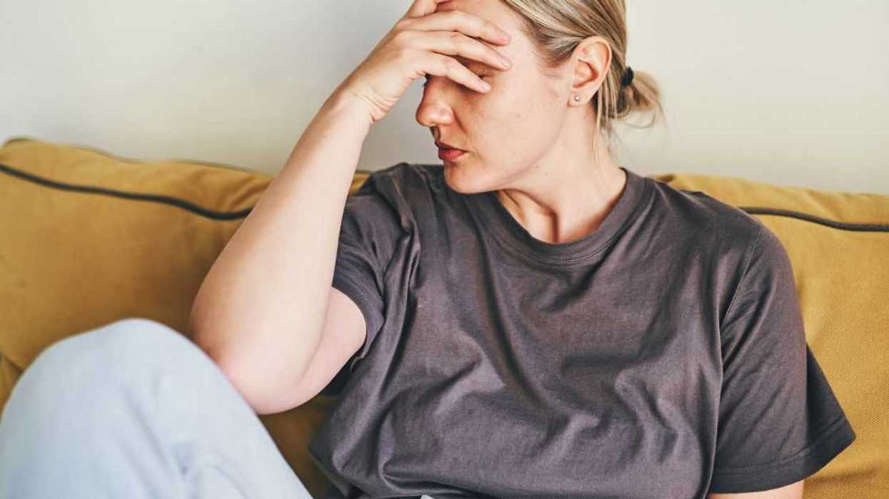 A young woman is sitting at home on a yellow sofa with her head in her hands, sad or showing a headache or frustration and depression.