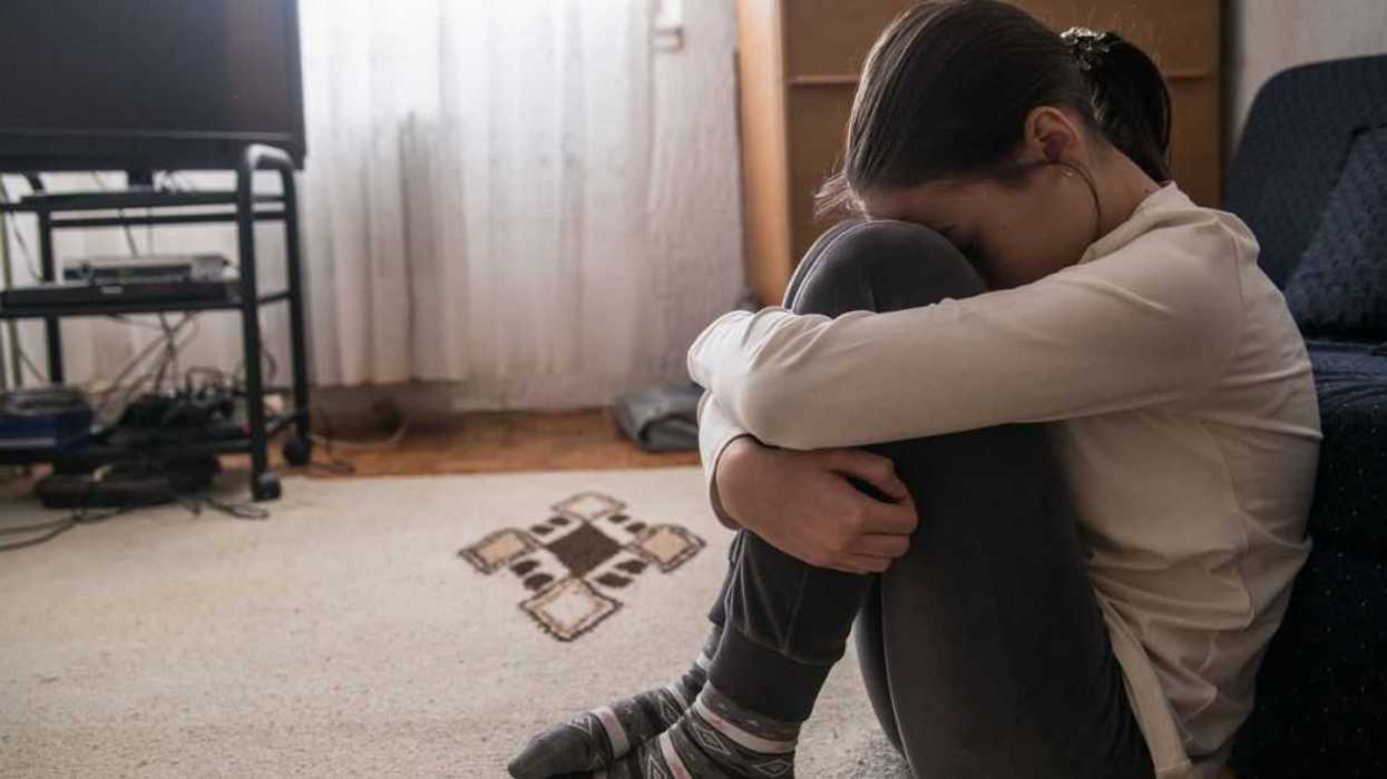 A young woman sits on the floor with her head down, holding her knees