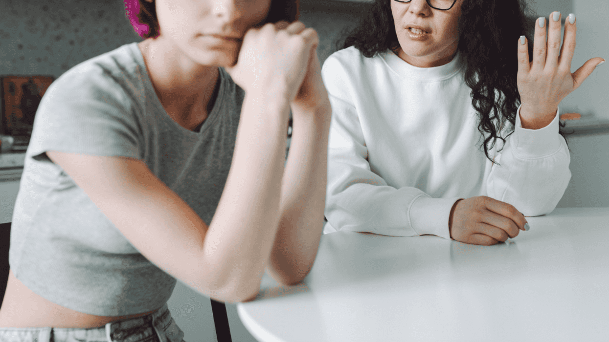 A young woman with pink hair sitting at a table with her hands crossed, and an older brown haired woman sitting behind her with one hand in the air.