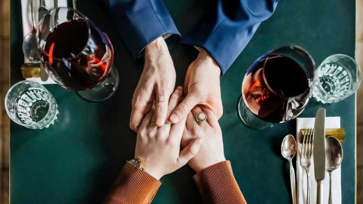 An aerial view of a couple holding hands and drinking red wine while sitting at a restaurant table for lunch together.