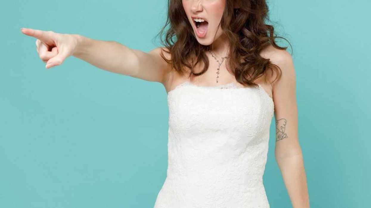 An angry, irritated bride young woman in beautiful white wedding dress points her index finger while side screaming while standing isolated on blue turquoise background; studio portrait.