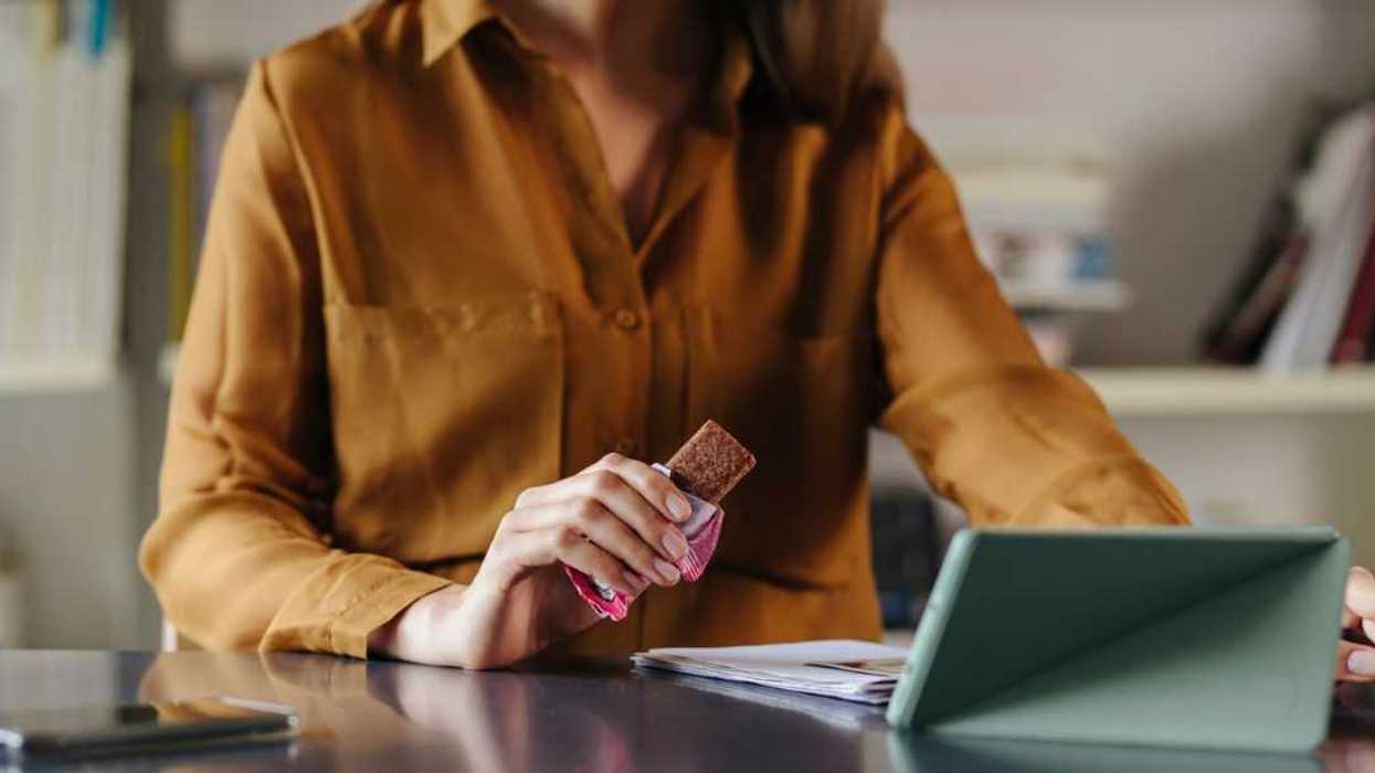 An anonymous businesswoman sitting at the office, eating a protein bar while video calling with her colleagues.