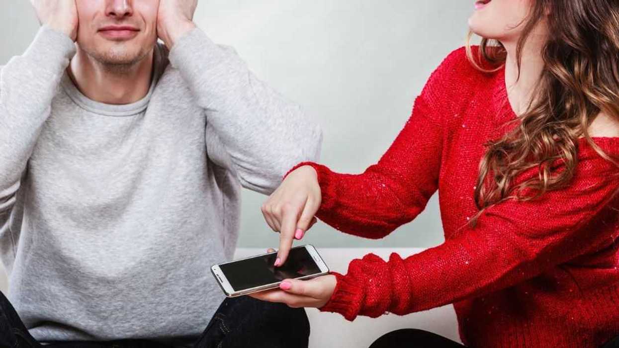 An outraged woman screams at her husband while pointing at his phone. He just sits and covers his ears.