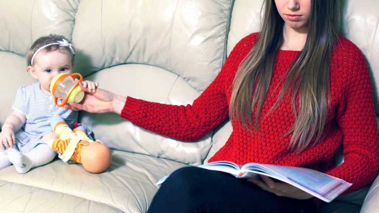 Babysitter in red sweater holds a bottle to a baby sitting next to her as she reads.