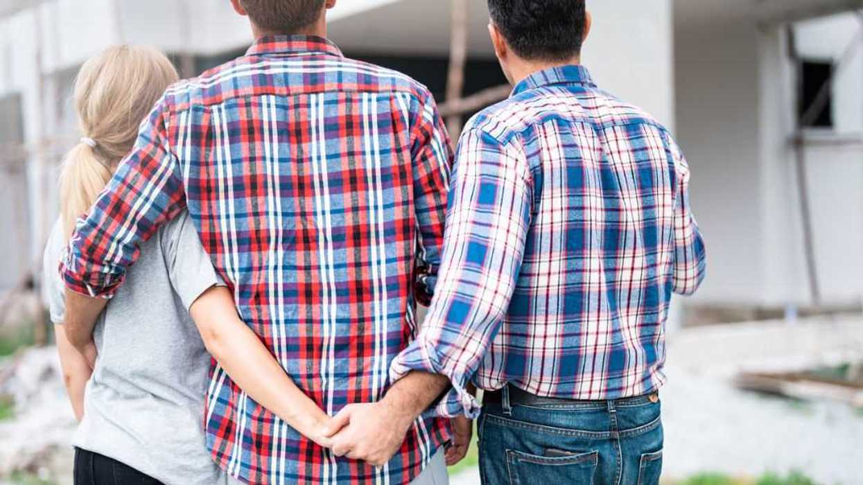 Back shot of a woman and two men. The man in the middle and the woman hold one another. She holds hands secretly with the other man.