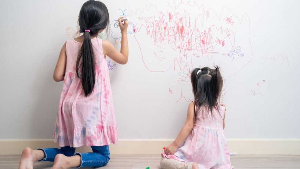 Back shot of two young girls drawing with markers on a white wall.