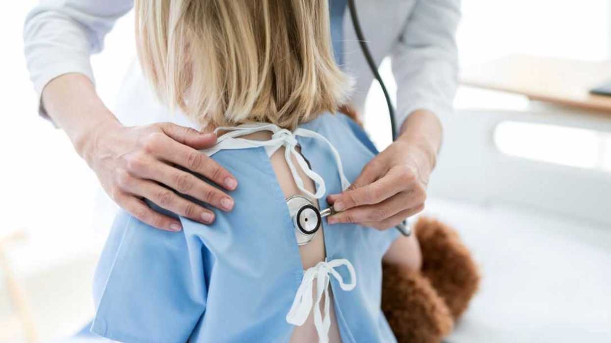 Backshot, cropped image of doctor examining girl with stethoscope in hospital. Ill child is wearing gown while sitting on bed. Healthcare worker is doing treatment of patient.