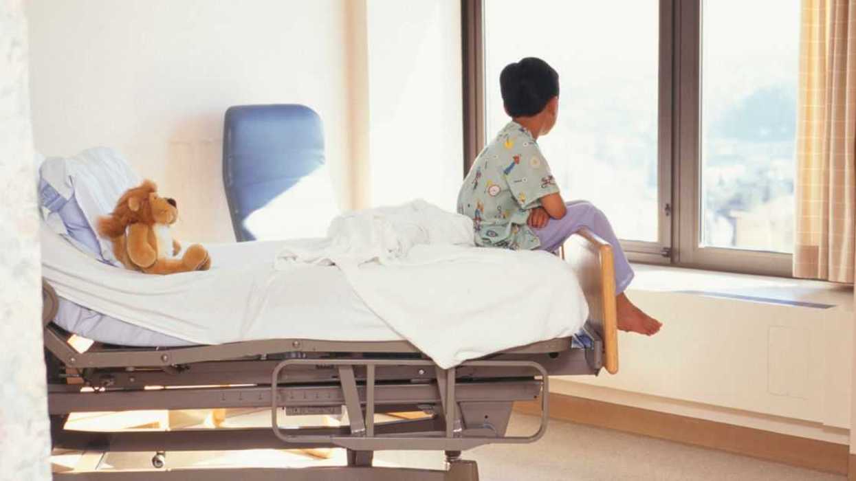 Backshot of a young boy (6-7) sitting on the edge of a hospital bed while gazing out the window.