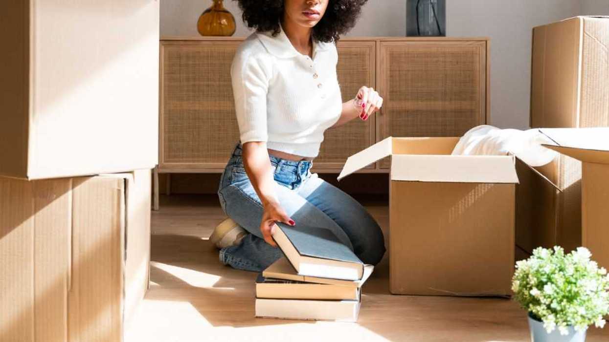 Beautiful woman unpacking her favorite books in her new apartment. Kneeling on the floor taking books out of cardboard boxes to place on the living room shelf.