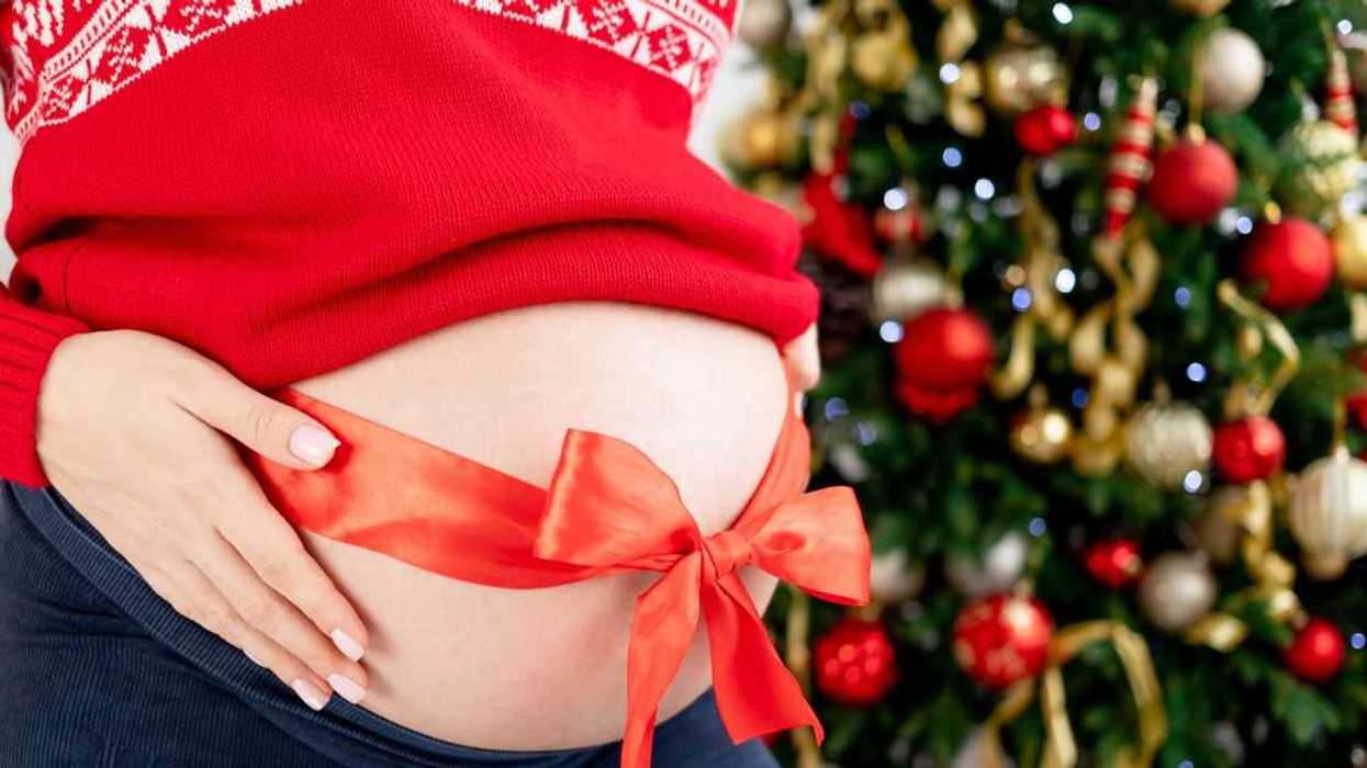 Belly of a pregnant woman with a red bow wrapped around her stomach, in a red sweater near the Christmas tree, close-up.
