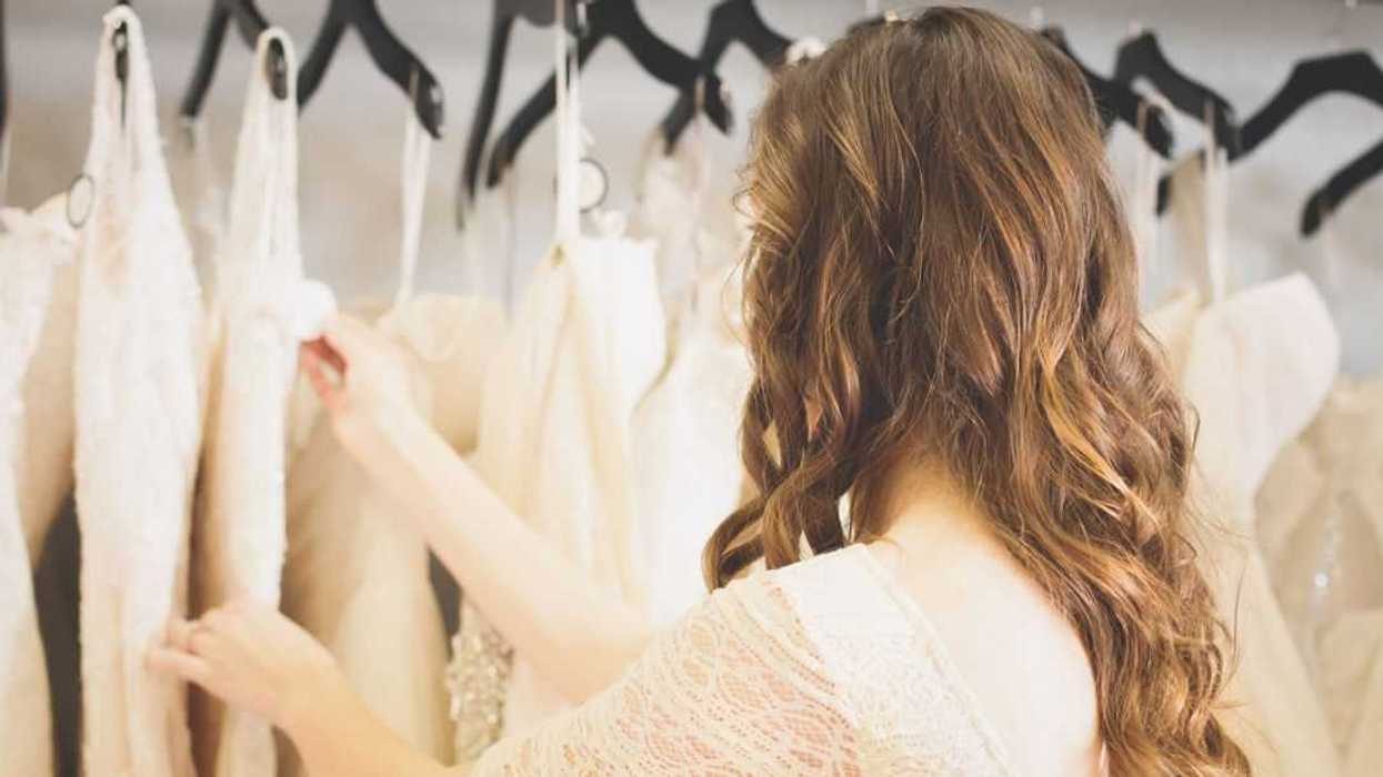 Bride-to-be shopping for wedding dress; head shot; facing dresses hanging on wall; fingering one in particular.