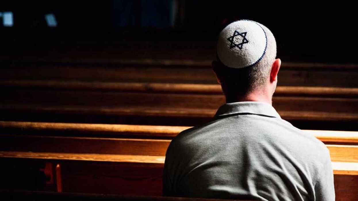 Close up, back-shot image depicting a young caucasian Jewish adult man in his 30s inside a synagogue. He has his head bowed in prayer and he is wearing the traditional Jewish skull cap - otherwise known as a kippah or yarmulke - on his head. The man has a beard and the background of the synagogue is blurred out of focus. Horizontal color image with copy space.