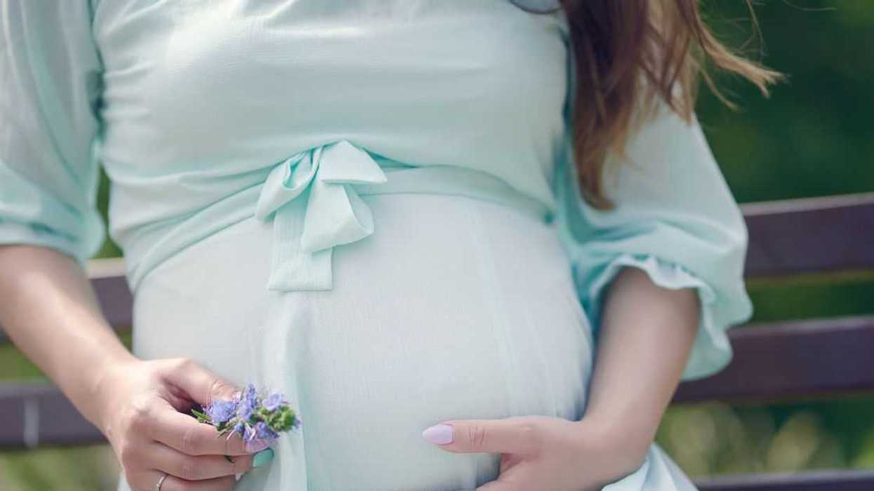 Close up of beautiful young woman, sitting on a bench during the day. She is in her last trimester of pregnancy, wearing a mint color, vaporous summer dress and holding flowers in her hands in a caressing gesture around her baby bump.