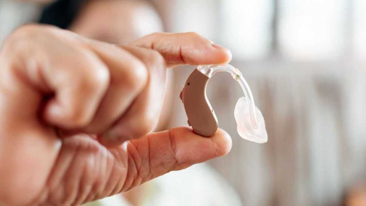 Close-up of senior hand holding a behind-the-ear hearing aid indoors.