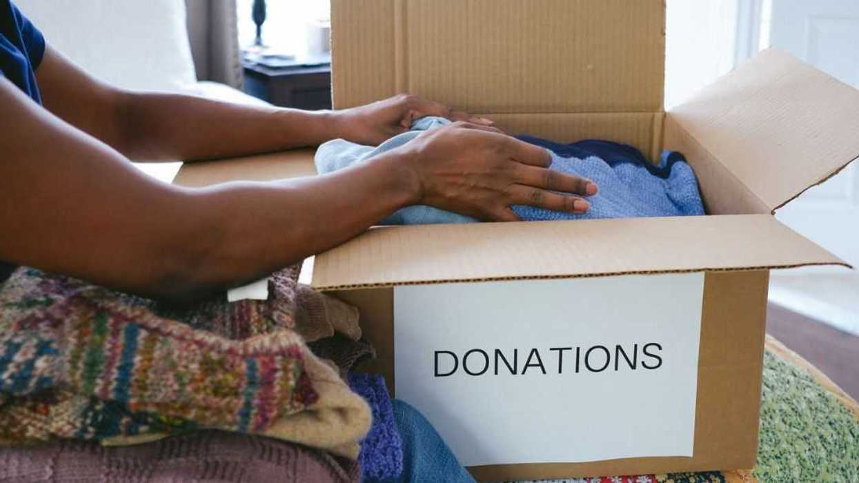 Close-up of unrecognizable woman sitting on bed packing box of clothes for donation.
