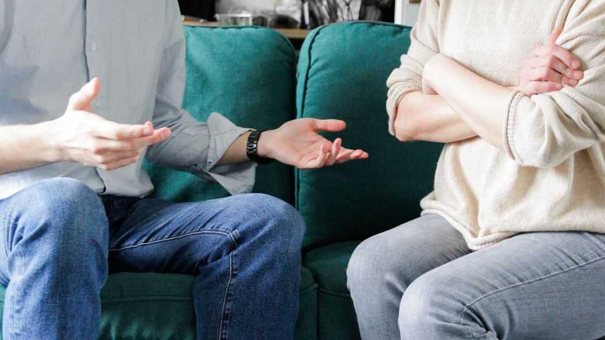 Close-up shot of a couple in a disagreement. His arms are open, hers are folded. They sit on a couch.