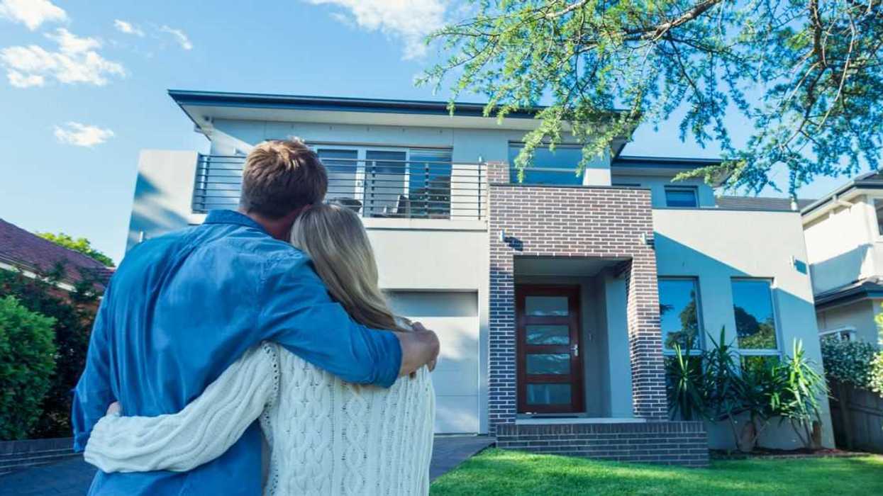 Couple standing in front of their new home. They are both wearing casual clothes and embracing. Rear view from behind them. The house is contemporary with a brick facade, driveway, balcony and a green lawn. The front door is also visible.