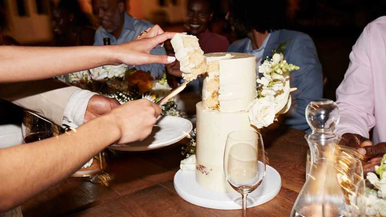 Cropped hands of newlywed couple cutting wedding cake near guests sitting at illuminated dining table during reception dinner party.