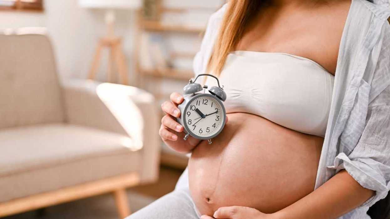 Cropped image, A pregnant woman in comfortable clothes sits on a chair in her living room, holding her belly and an alarm clock. Awaiting her baby's due date.